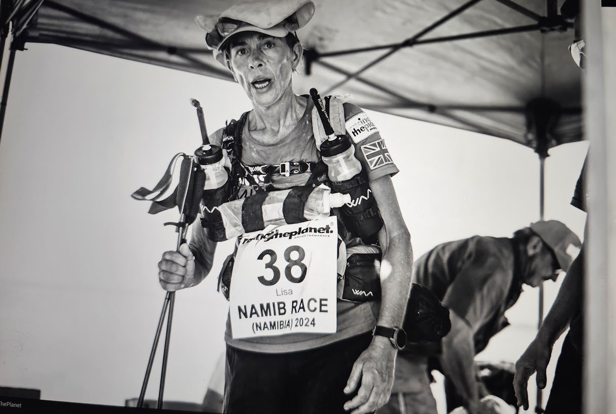 Lisa mid-stage at RTP Namibia 2024, sweat-stained, exhausted, race bib 38, in 50°C heat under a race tent