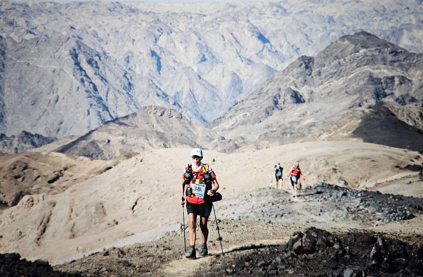 Lisa climbing a mountain trail with poles, race bib 38, in Namibia with hazy mountain ridges behind